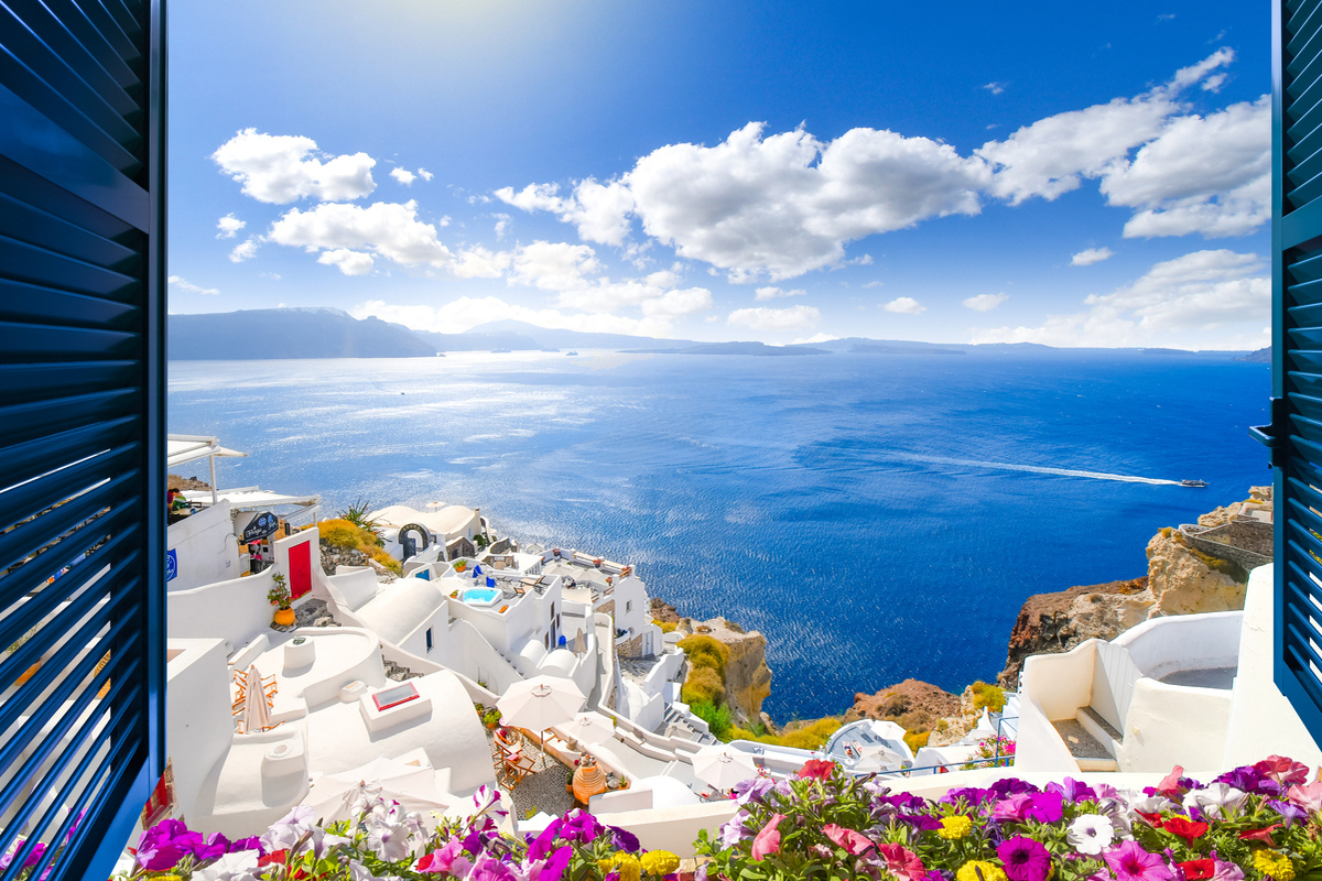 Widok przez otwarte okno z okiennicami na wybieloną wioskę Oia, wznoszącą się nad błękitnym Morzem Egejskim i kalderą na wyspie Santorini w Grecji.